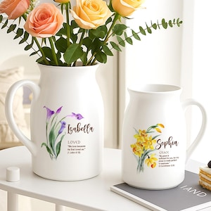 May include: Two white ceramic jugs with handles, decorated with floral designs and names. One has purple calla lilies and the name "Isabella", the other yellow daffodils and the name "Sophia".