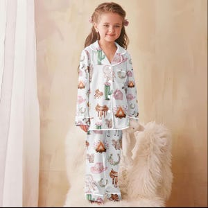 Puede incluir: Conjunto de pijama blanco para ni&ntilde;os con estampado de tem&aacute;tica Western. El estampado incluye cactus, sombreros de vaquero, sillas de montar y hogueras. La parte superior de manga larga y los pantalones tienen ribetes blancos.