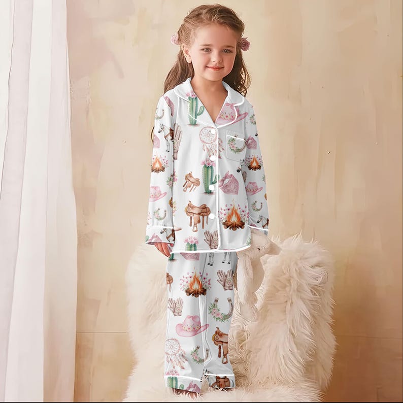 Puede incluir: Conjunto de pijama blanco para ni&ntilde;os con estampado de tem&aacute;tica Western. El estampado incluye cactus, sombreros de vaquero, sillas de montar y hogueras. La parte superior de manga larga y los pantalones tienen ribetes blancos.