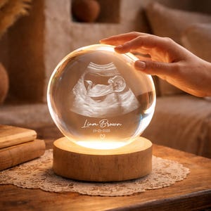 Peut inclure: Une sphère en verre transparent avec une image échographique d'un bébé, posée sur une base en bois. La sphère est éclairée de l'intérieur, affichant le nom "Liam Brown" et la date "01-23-2026" avec un symbole de cœur. La sphère est sur une table en bois.