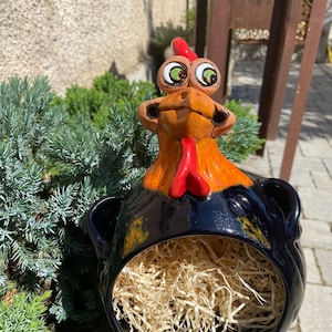 Könnte beinhalten: Ein schwarzer Keramikhahn mit orangefarbenen und roten Akzenten. Der Hahn hat große, grüne Augen und einen breiten, offenen Schnabel. Der Körper des Hahns ist hohl und mit Stroh gefüllt.