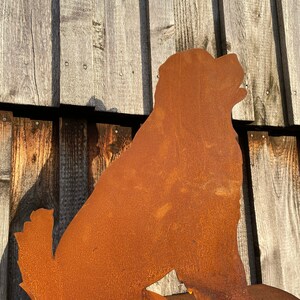 Könnte beinhalten: Eine rostige Metallsilhouette eines sitzenden Hundes, der den Kopf zur Seite dreht. Der Hund blickt nach rechts und steht auf einem rostigen Metallsockel.