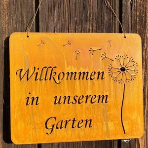 Könnte beinhalten: Ein rostiges Metallschild mit der Aufschrift "Willkommen in unserem Garten" in schwarzer Schrift. Das Schild ist mit Löwenzahnmustern verziert.