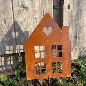 Könnte beinhalten: Ein rostiger, hausförmiger Gartenpfahl aus Metall mit einem Herz-Ausschnitt im Dach. Der Pfahl ist von grünen Pflanzen umgeben.