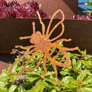 Könnte beinhalten: Eine rostige Metallbienenskulptur mit filigranen Details, die in einem Beet mit grünen Pflanzen steht.