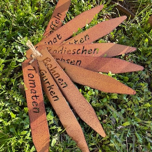 Könnte beinhalten: Ein Satz von zehn rostigen Gartenmarkierungen aus Metall mit verschiedenen Pflanzennamen darauf. Die Namen sind "Tomaten", "Gurken", "Bohnen", "Erbsen", "Radieschen", "Zwiebeln", "Salat", "Pfeffer" und zwei weitere, die auf dem Bild nicht zu sehen sind.
