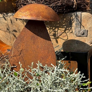 Könnte beinhalten: Eine rostige Metallpilzskulptur mit abgerundeter Oberseite und einer tränenförmigen Basis. Die Skulptur ist von einem Bett aus silbrig-grünem Laub umgeben.