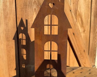 Könnte beinhalten: Ein rostiges, hausförmiges Metallausschnitt mit einem Herzen oben und drei Fenstern. Das Haus steht auf einer Holzoberfläche.