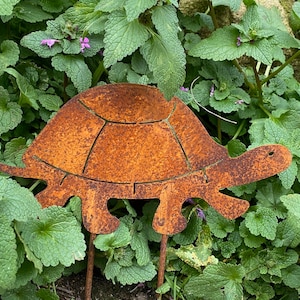 Könnte beinhalten: Ein rostiger Metall-Schildkröten-Gartenpfahl mit einem detaillierten Panzerdesign. Der Pfahl ist in einem Beet mit grünen, blättrigen Pflanzen platziert.