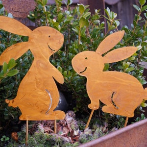 Könnte beinhalten: Zwei rostige Metallhasen mit lächelnden Gesichtern stehen in einem mit grünen Pflanzen gefüllten Topf.