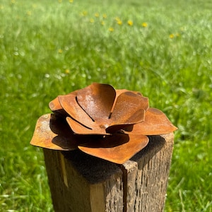 Könnte beinhalten: Eine rostige Metallblumenskulptur auf einem verwitterten Holzpfosten. Die Blüte hat überlappende Blütenblätter, die einen dreidimensionalen Effekt erzeugen. Der Hintergrund zeigt eine grüne Wiese und einen blauen Himmel.