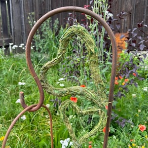 Könnte beinhalten: Ein rostiger Metallgartenbogen mit zwei herzförmigen Kränzen aus getrocknetem Moos und Zweigen. Der Bogen ist von grünem Gras und Wildblumen umgeben.