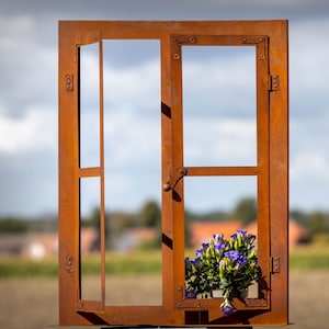 Könnte beinhalten: Eine verwitterte, rostige Metallfensterrahmen-Skulptur mit einem offenen Fenster. Der Rahmen hält einen kleinen Pflanzkasten mit leuchtend violetten Blumen. Der Hintergrund zeigt eine verschwommene Landschaft unter einem bewölkten Himmel.