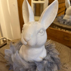 Könnte beinhalten: Eine weiße Keramik-Hasenkopf-Figur mit langen Ohren, umgeben von einem Kranz aus grauen Federn. Der Hase hat eine glänzende Oberfläche und detaillierte Gesichtszüge. Ein dekorativer Artikel.