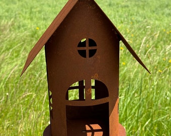 Könnte beinhalten: Ein rostiger, hausförmiger Kerzenhalter aus Metall mit ausgeschnittenen Fenstern und einem abgerundeten Dach. Der Halter ist auf einem Holzpfosten montiert.
