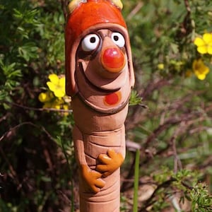 Könnte beinhalten: Ein skurriler Keramik-Gartenzwerg mit roter Mütze, einer großen roten Nase und einem breiten Lächeln. Der Zwerg steht in einer Gartenlandschaft mit grünen Pflanzen und gelben Blumen.