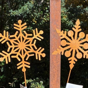 Könnte beinhalten: Zwei rostige Metall-Schneeflocken-Gartenornamente mit filigranen Designs. Die Schneeflocken sind an Metallpfählen befestigt.