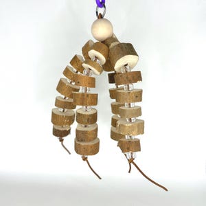 May include: A bird toy made of multiple strands of small, round wooden pieces threaded with clear beads. The toy hangs from a purple chain and a wooden bead. The wooden pieces are light brown and have a natural, textured appearance.