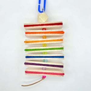 May include: A rainbow-coloured wooden toy with a blue clip and a wooden bead at the top. The toy is made of multiple wooden pieces with different coloured felt strips. The toy is designed for small animals to play with.