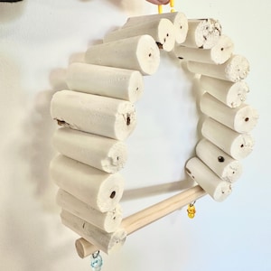 May include: A circular bird perch constructed from light-coloured wooden dowels. The perch features a wooden dowel for the bird to sit on, and a small bell suspended from it. Designed for hanging in a bird cage.