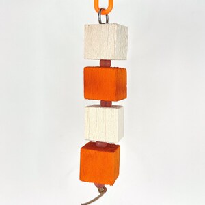 Può includere: Un giocattolo di legno con tre blocchi impilati. I blocchi superiore e inferiore sono bianchi, e il blocco intermedio è arancione. I blocchi sono collegati da perline rosa.