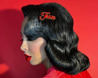 May include: A woman with dark hair styled in waves wears a red velvet robe with a fur trim. She has red lipstick and a hair clip that says "Tease" in red letters.