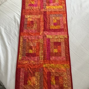 May include: A rectangular, handmade quilt in shades of red, orange, and yellow. The quilt features a log cabin design with squares and rectangles. The quilt has a red border and red hanging loops at the top.