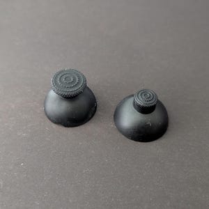 May include: Two black joystick thumbsticks with a textured top and a rounded base. The thumbsticks are designed for a gaming controller and are made of a matte black material. They are shown on a dark gray surface.