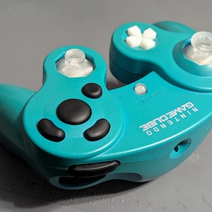 Puede incluir: Mando Nintendo GameCube de color turquesa con botones negros y cruceta blanca. El mando tiene dos palancas analógicas y el logotipo de Nintendo GameCube es visible.