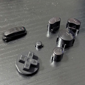 Puede incluir: Un conjunto de botones negros de mando de videojuegos, que incluye una cruceta, un botón Z y varios botones con letras. Los botones están dispuestos sobre una superficie oscura, probablemente para reparación o personalización.