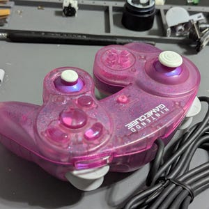 Könnte beinhalten: Ein transparent rosa Nintendo GameCube Controller mit Glitzer-Finish. Der Controller hat einen weißen Joystick und Knöpfe sowie ein schwarzes Kabel. Die Worte "NINTENDO GAMECUBE" sind auf dem Controller aufgedruckt.