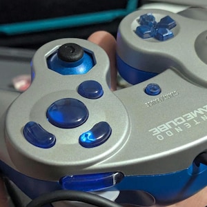 Może przedstawiać: Srebrno-niebieski kontroler Nintendo GameCube. Kontroler ma szarą obudowę z niebieskimi akcentami i przyciskami. Na kontrolerze wydrukowano tekst "NINTENDO GAMECUBE".