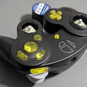 May include: Black video game controller with yellow buttons and a white logo. The controller has a unique design with two circular sections and a curved handle. A small battery is visible in the background.