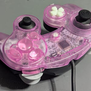 Può includere: Un controller per videogiochi rosa traslucido con joystick e pulsanti neri. Il controller ha un guscio di plastica trasparente, che rivela i componenti interni. Sono visibili pulsanti bianchi e rosa. Un cavo nero si estende dal basso.