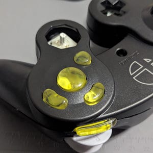 May include: Black video game controller with yellow buttons and a white logo. The controller has a distinctive shape with a curved handle and a circular button layout. The yellow buttons are translucent and the controller is partially disassembled.