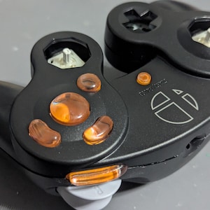 Puede incluir: Mando de videojuegos negro con botones y detalles en naranja. El mando tiene una cruceta, botones y un joystick. Un logotipo y texto blancos son visibles en la superficie.