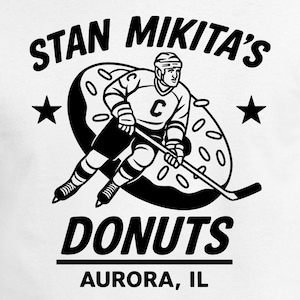 May include: White t-shirt featuring a black and white graphic of a hockey player skating through a donut. The text reads "STAN MIKITA'S DONUTS AURORA, IL" with star accents.
