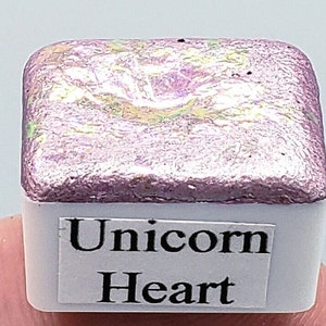 Pode incluir: Uma pequena panela quadrada branca de tinta aquarela rosa iridescente com as palavras "Unicorn Heart" impressas no rótulo.
