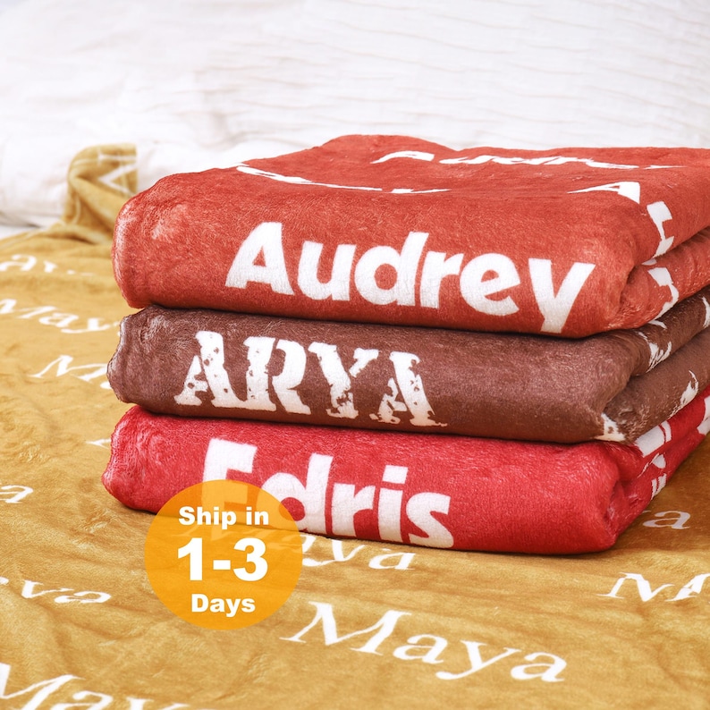 May include: A stack of three folded plush blankets in rust, brown, and red, each featuring the white printed names Audrey, ARYA, and Edris. Beneath the stack lies a gold-colored blanket with the repeating white printed name Maya.