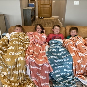 May include: Four soft plush blankets in mustard yellow, dusty rose, teal, and burnt orange, each featuring a repeating white name pattern. The blankets are draped over four children sitting on a tan sofa, displaying the names Case, Winnie, Maverick, and Huck.