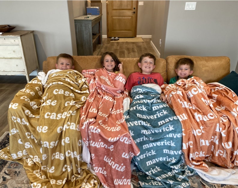 May include: Four soft plush blankets in mustard yellow, dusty rose, teal, and burnt orange, each featuring a repeating white name pattern. The blankets are draped over four children sitting on a tan sofa, displaying the names Case, Winnie, Maverick, and Huck.