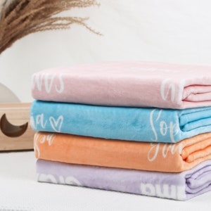May include: A stack of four folded plush blankets in pastel pink, light blue, orange, and lavender. Each blanket features white cursive text and a heart graphic printed along the folded edge, showcasing a soft, fuzzy texture throughout the pile.