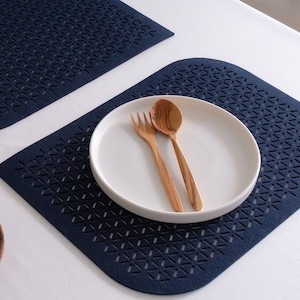 May include: Two blue placemats with a geometric pattern, one rectangular and one oval, with a white plate, wooden spoon, and fork on top.