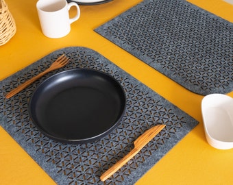 Op de afbeelding: Een zwart bord met een houten vork en mes op een grijze placemat met een geometrisch patroon. De placemat ligt op een geel oppervlak.