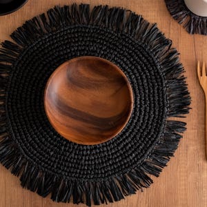 May include: Black woven placemat with a fringe border and a wooden bowl in the center. The placemat is on a wooden table.
