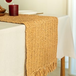 Peut inclure: Un chemin de table en osier de couleur beige avec des franges sur les bords. Le chemin de table est posé sur une nappe blanche et une table en bois.