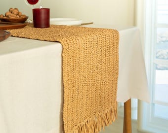 May include: A tan woven table runner with fringe edges. The runner is on a white tablecloth and a wooden table.