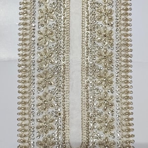 以下が含まれることがあります： 花と真珠のようなビーズ装飾が施された、詳細な金色の刺繍入りネックライン。対称的な花の模様と中央の白い布パネルが特徴です。複雑なビーズ細工が、豪華で装飾的な要素を作り出しています。