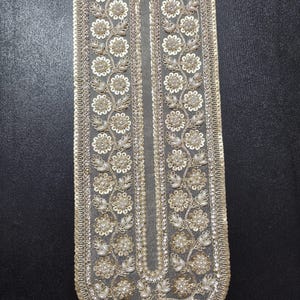 May include: A decorative, elongated textile piece with intricate floral embroidery. The design features white flowers, pearls, and sequins on a gray background, bordered with a gold trim. The piece is likely for embellishment or apparel.