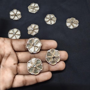 Può includere: Una collezione di piccole toppe circolari a forma di fiore con tessuto a rete color oro metallizzato, dettagli in filo d'oro ricamato e un accento centrale in strass trasparente, disposte su una superficie scura e in un palmo.